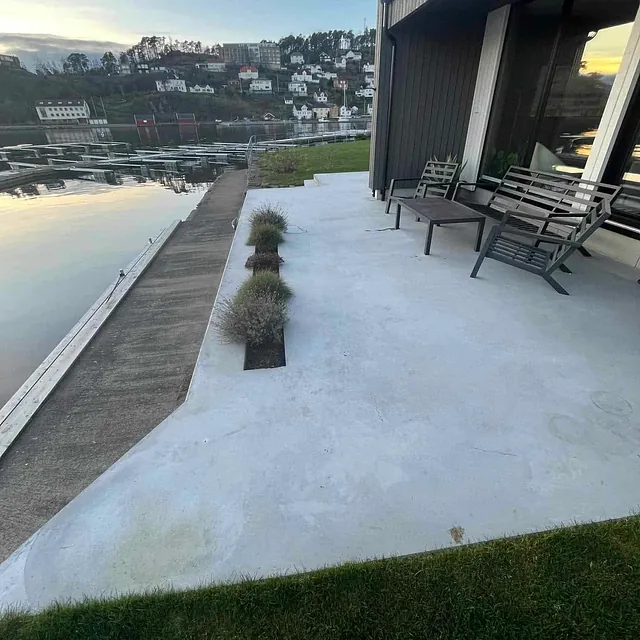 Betongterrasse med utemøbler ved moderne hus, med utsikt over vann og småhusbebyggelse.