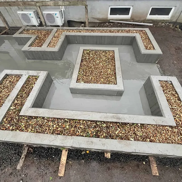 Rektangulære plantekasser i betong fylt med bark, plassert i mønster på våt betongflate.