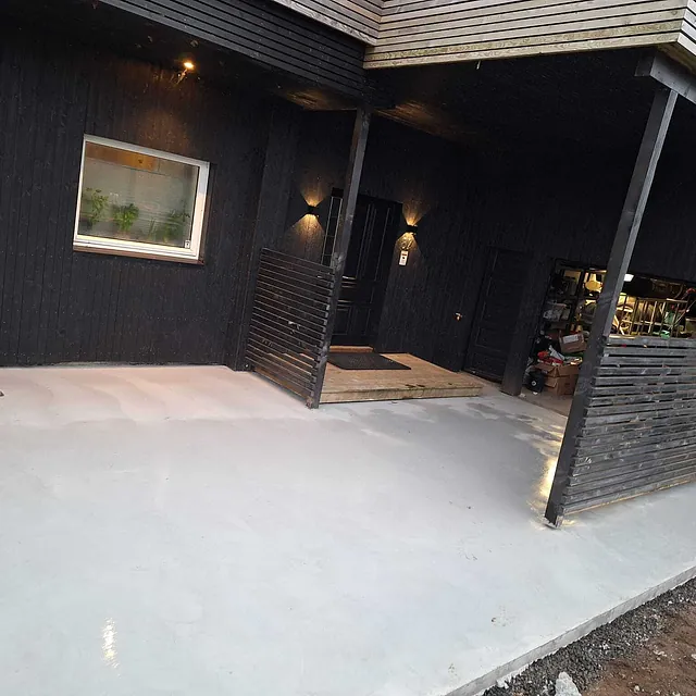 Ny betongplatting foran inngangsparti på hus med mørk trekledning og overbygd tak.