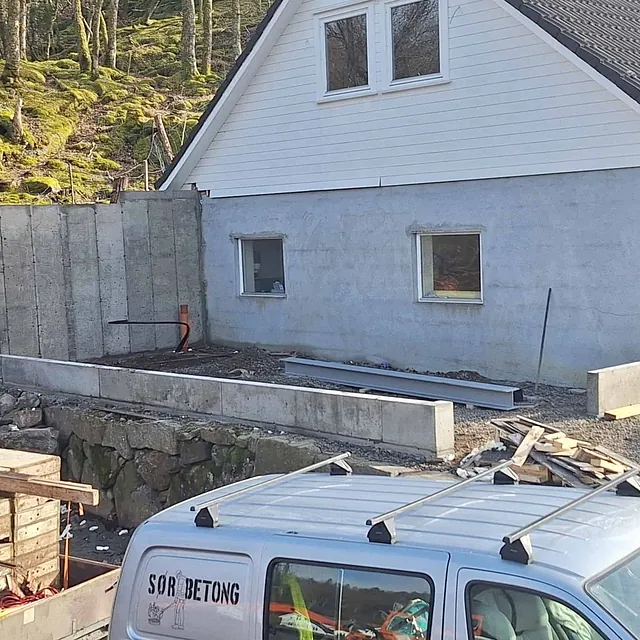 Delvis ferdig betongmur ved hus med skog i bakgrunnen og byggeutstyr foran.