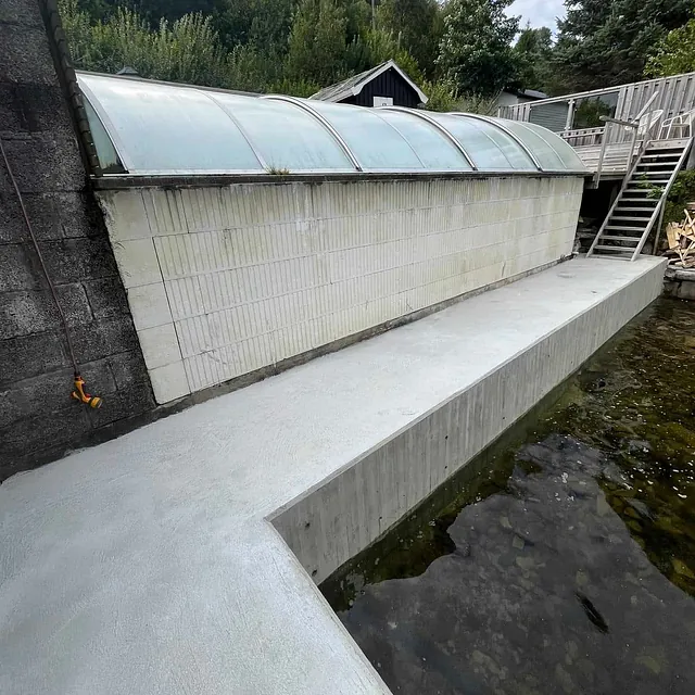 Betongdekke langs bygning ved vannkant, omgitt av skog og treterrasse med trapp.