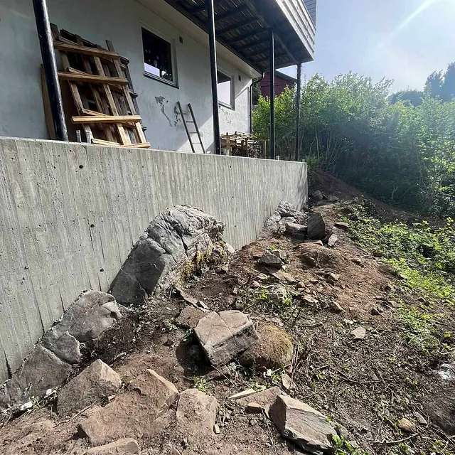 Støttemur i betong ved hus med skrånende terreng og synlig fjell og jord.