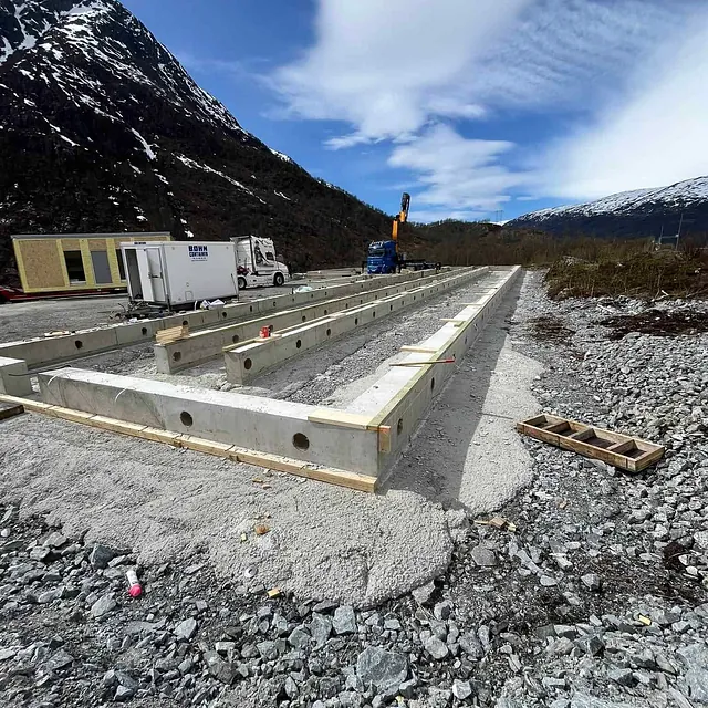 Byggeplass med betongfundament i fjellterreng, omgitt av lastebiler og kran.