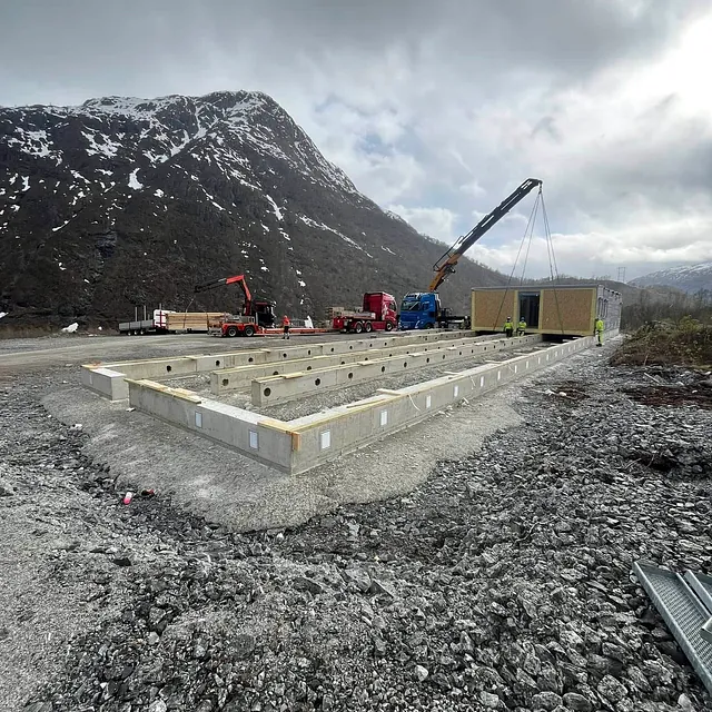 Kran løfter bygningsmodul på betongfundament i fjellandskap med snødekte fjell i bakgrunnen.